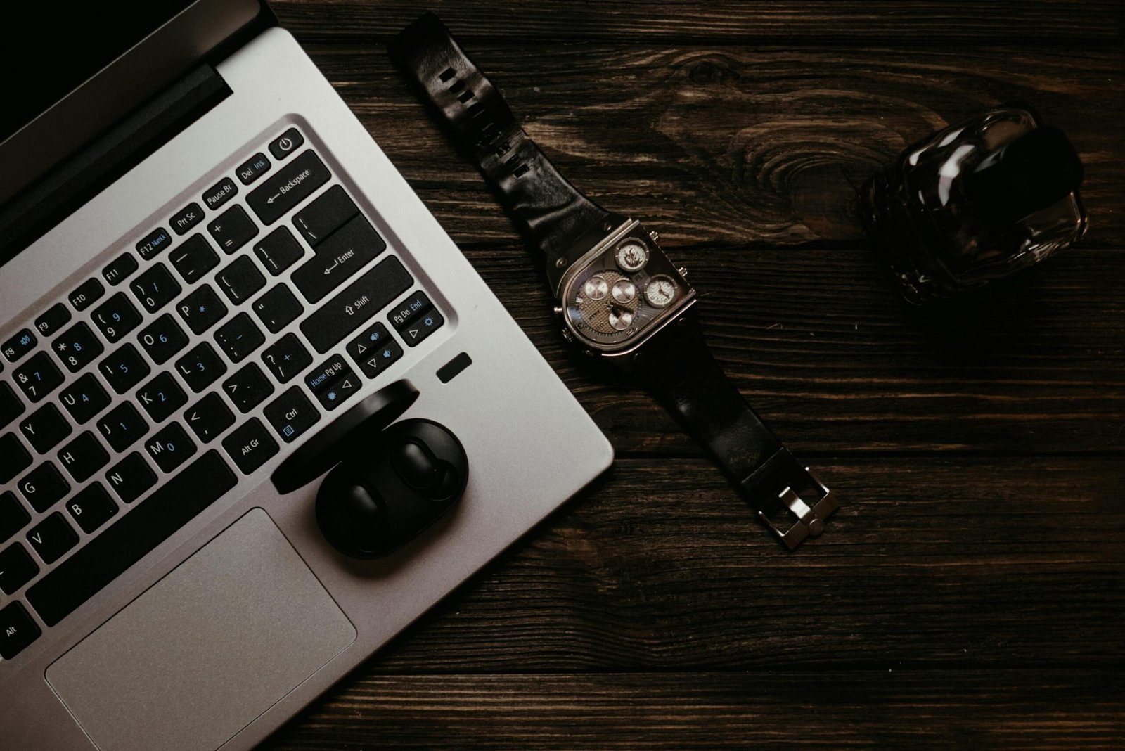 A stylish flat lay featuring a laptop, leather watch, earphones, and a perfume bottle on a dark wooden table.